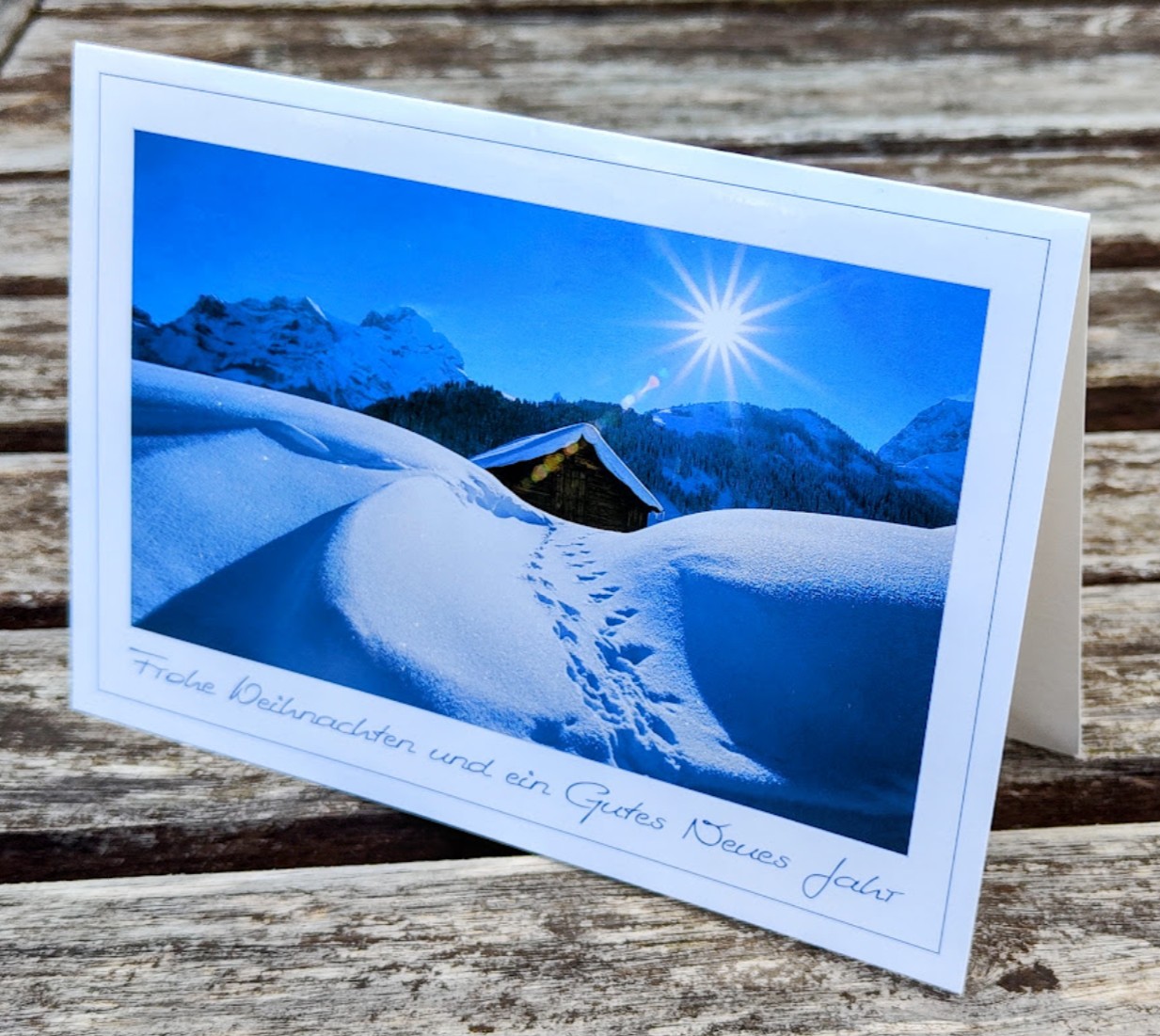 Abgebildet ist eine Berghütte in einer Winterlandschaft. Folgender Text steht auf der Karte: Frohe Weihnachten und ein Gutes Neues Jahr